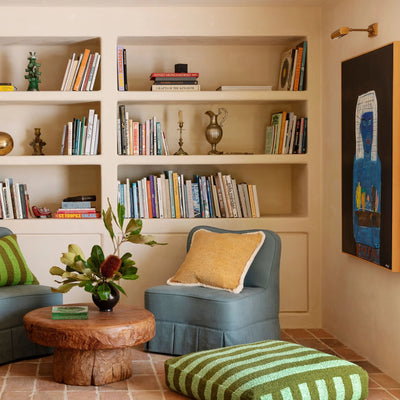 A cozy reading nook with built-in bookshelves, two blue chairs, a wooden coffee table, and a green striped pouf. A painting hangs on the wall, adding an artistic touch.