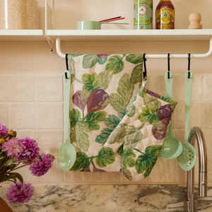 Fig-patterned kitchen towel and figgy green quilted linen oven mitt hanging with mint green utensils above a marble countertop.