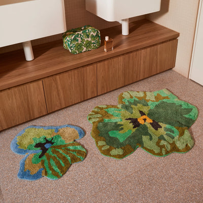 green pansy and viola green small bath mats placed on a tiled bathroom floor in front of a wooden vanity with a  green floral toiletry bag and perfume bottle.