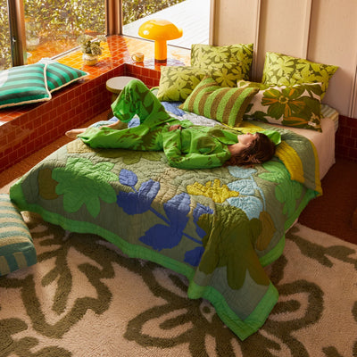 Woman wearing green floral pajama lying in the bead with floral quilted throw and stripe cushion and pillow with printed floral pillowcase
