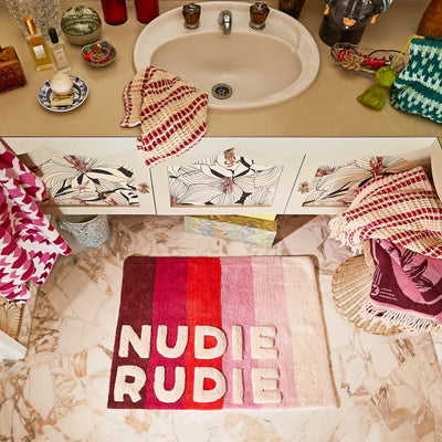 colourful bathroom with a striped mat that says “NUDIE RUDIE.” The sink area has fun patterns, and towels