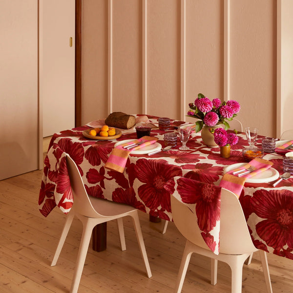 Dining table set with a red floral tablecloth, pink napkins, and a vase of bright pink flowers.