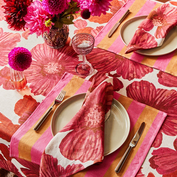 Table setting with red floral tablecloth, leon cerise linen napkins set of four, striped pink and gold placemats, and pink flowers in vases.
