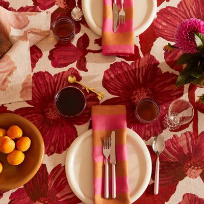 Table set with leon cerise round tablecloth, striped pink and gold napkins, white plates, and drinks, with a bowl of apricots.