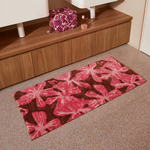 mave pink bath mats placed on a tiled bathroom floor in front of a wooden vanity with a floral toiletry bag and perfume bottle.