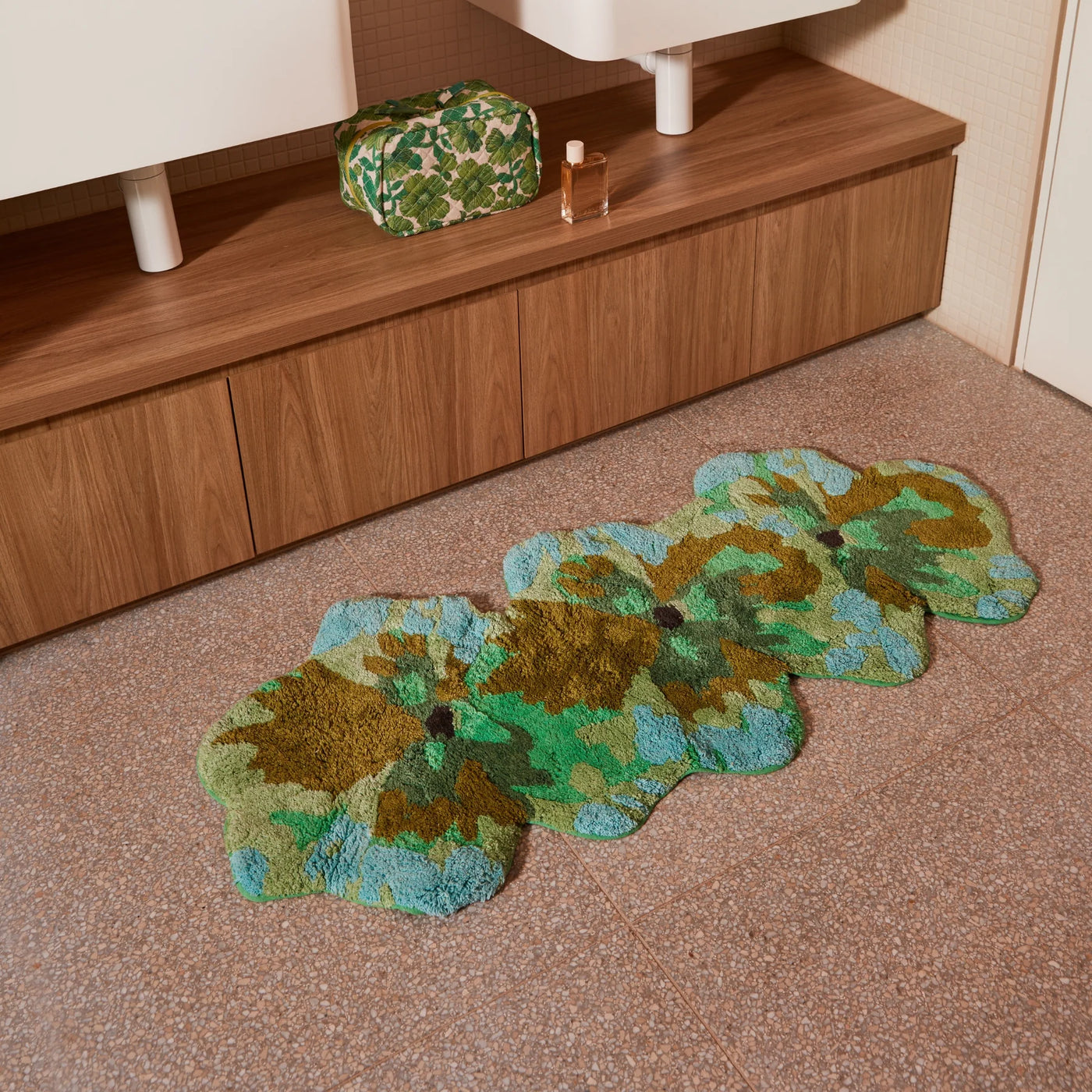 pansy long bath mat placed on a tiled bathroom floor in front of a wooden vanity with a floral toiletry bag and perfume bottle.