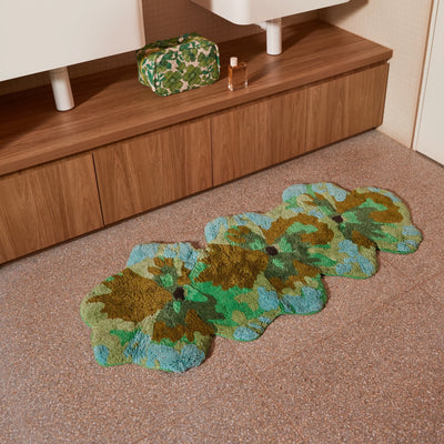 pansy long bath mat placed on a tiled bathroom floor in front of a wooden vanity with a floral toiletry bag and perfume bottle.