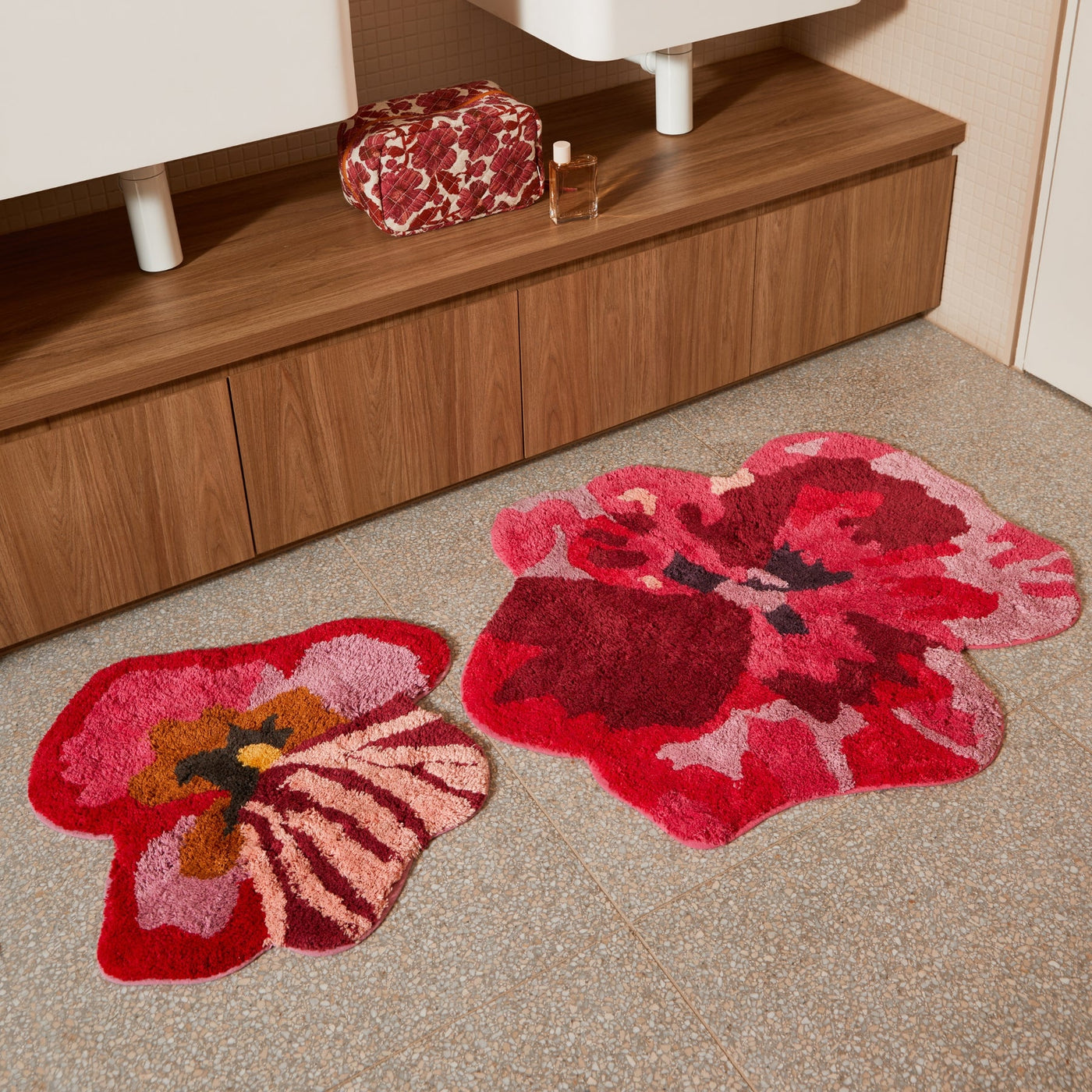 viola pink small bath mat and pansy bath mat placed on a tiled bathroom floor in front of a wooden vanity with a floral toiletry bag and perfume bottle.