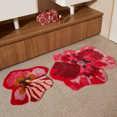 viola pink small bath mat and pansy bath mat placed on a tiled bathroom floor in front of a wooden vanity with a floral toiletry bag and perfume bottle.