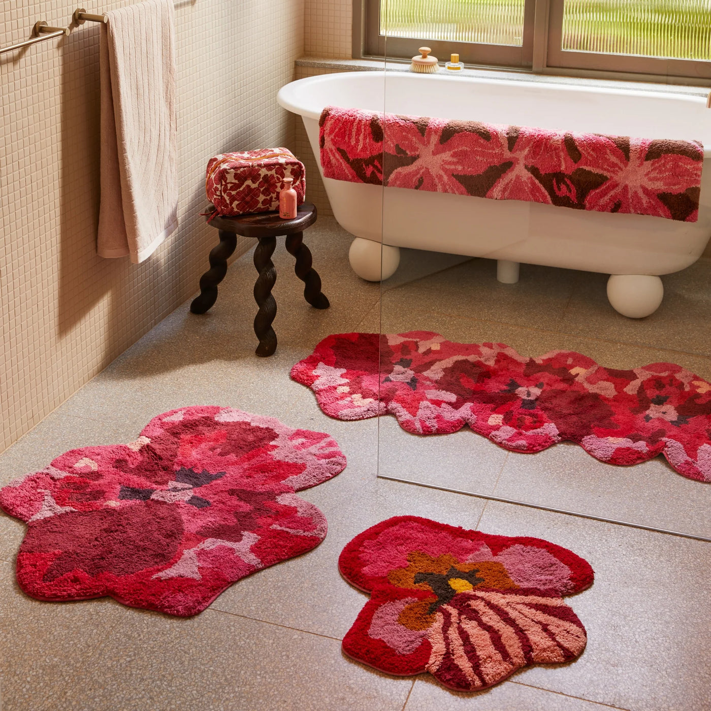 Bathroom with a white freestanding bathtub, pansy bath mat, viola pink small bath mat and  pink and red floral bath mats on the floor, and a matching floral towel draped over the tub.