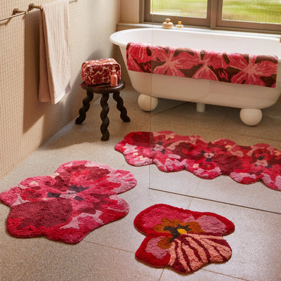 Bathroom with a white freestanding bathtub, pansy bath mat, viola pink small bath mat and  pink and red floral bath mats on the floor, and a matching floral towel draped over the tub.