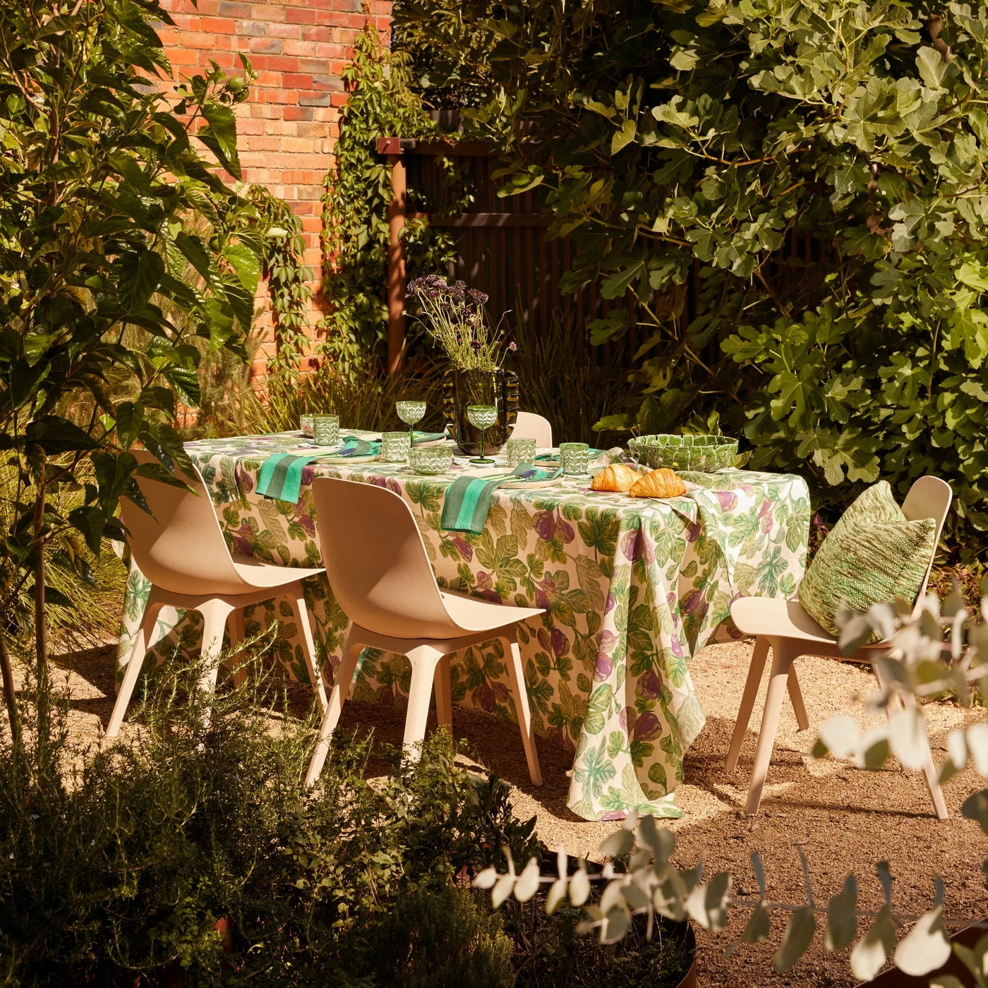 Outdoor dining setup with a fig-patterned tablecloth, green tableware, and croissants in a lush garden setting.