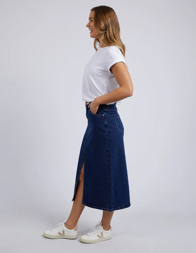 side of view of woman standing wearing foxwood scout midi skirt indigo, white top, and white shoes