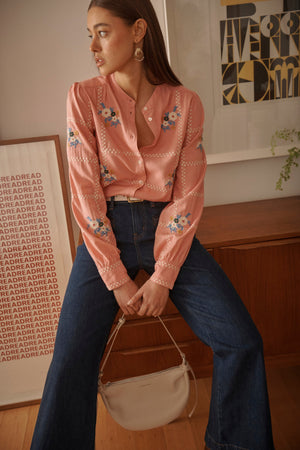 Kireina Carol Shirt in Musk Pink – model seated wearing the floral-embroidered musk pink button-up shirt with wide-leg jeans and tan shoulder bag, styled against a typographic art print backdrop