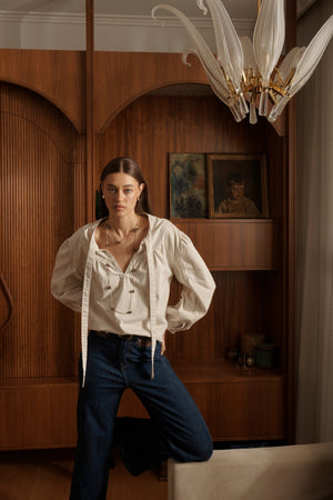 Campaign shot of Kelly Shirt Sand with open pussybow neckline and puffed sleeves styled with dark jeans in elegant interior setting