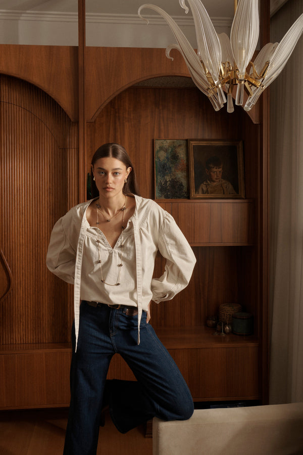 Campaign shot of Kelly Shirt Sand with open pussybow neckline and puffed sleeves styled with dark jeans in elegant interior setting